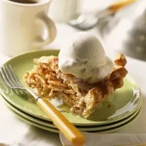 Fotografia de um sorvete de canela sobre uma torta de massa folhada recheada com maçã, que está ao lado de um garfo. A sobremesa está sobre três pratos verdes empilhados, que estão apoiados sobre uma toalha de mesa branca.
