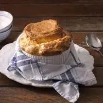 Fotografia em tons marrom de bancada de madeira, um prato com um pano listrado em azul por cima, no prato um pote com o suflê, do lado esquerdo uma colher e no direito dois potes brancos.