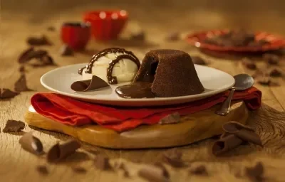 Fotografia em tons de marrom em uma bancada de madeira marrom. Ao centro, uma tábua de madeira, com um pano vermelho e um prato branco em cima contendo o petit gateau. Ao redor, há alguns pedaços de chocolates espalhados. Fotografia em tons de marrom em uma bancada de madeira marrom. Ao centro, uma tábua de madeira, com um pano vermelho e um prato branco em cima contendo o petit gateau. Ao redor, há alguns pedaços de chocolates espalhados.