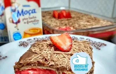 Imagem de um pedaço da receita de Pavê em camadas, servido em um prato, com cobertura de chocolate e decorado com um morango. Ao fundo vê-se embalagens do produto Moça Pra Toda a Família e a travessa contendo o restante da receita.