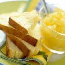 Fotografia em tons de amarelo, em uma bancada de madeira de cor branca. Ao centro, um prato verde contendo as fatias de pão com um recipiente ao lado contendo alguns pedaços de frutas.