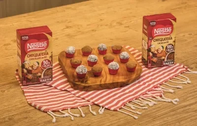 Fotografia em tons de vermelho em uma bancada de madeira de cor marrom. Ao centro, um pano branco com listras vermelhas com uma tábua de madeira em cima contendo os brigadeiros. Ao lado, há duas caixinhas com cacau e chocolate em pó dois frades.