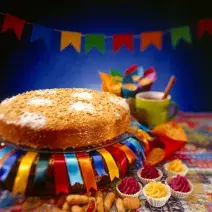 Fotografia em tons coloridos em uma bancada com uma toalha junina, um bolo redondo em cima de um prato com fitas de cetim decorando ele e bandeirolas de festa junina ao fundo.