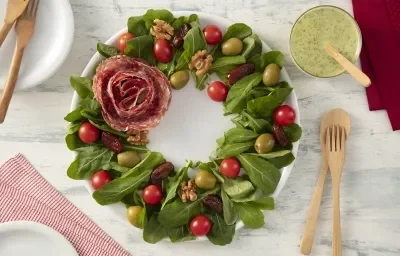 Foto da receita de Salada Guirlanda de Natal. Observa-se a rúcula, os tomates, a azeitona e as tâmaras em formato de guirlanda no prato branco, enquanto o salame está em formato de rosa ao lado esquerdo.