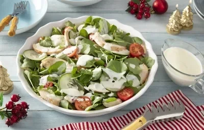 Foto em tons claros da receita de salada de chester com molho de iogurte servida em um bowl grande de porcelana branca com uma xícara de iogurte ao lado sobre uma mesa de madeira azul