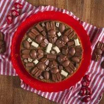 Fotografia em tons de vermelho, em uma bancada de madeira de cor marrom. Ao centro, uma toalha vermelha listrada com um prato vermelho em cima contendo o cookie em fatias. Ao redor do prato, há alguns KitKat's bunny e mini ovinhos de KitKat.