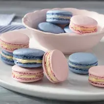 Fotografia em tons de azul e rosa em uma bancada de madeira branca clara com vários macarons coloridos em um prato branco e um paninho azul listrado.