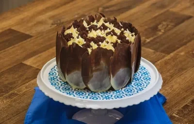 Foto aproximada de um bolo de chocolate com decoração de lascas marmorizadas, sobre um prato branco e azul em uma bancada de madeira