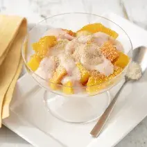 Fotografia em tons de branco e amarelo de uma bancada branca com um prato retangular branco, sobre ele uma taça com a salada de frutas e uma colher. Ao lado um paninho amarelo.