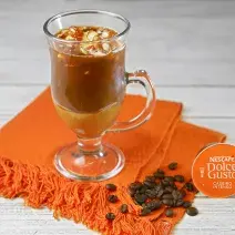Fotografia em tons de laranja em uma bancada de madeira branca, um pano laranja, uma xícara de vidro com o café com sorvete e pé de moleque dentro dele. Ao lado, uma cápsula de café caseiro intenso Dolce Gusto.