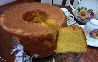 Foto da receita de bolo de milho com leite moça servida em uma porção grande com uma fatia cortada sobre um base de vidro circular com canecas e bules decorados ao fundo