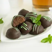 Fotografia em tons de branco, marrom e verde de uma bancada branca vista de frente, contém um prato branco com bombons de chocolate e folhas de hortelã em volta para decorar.