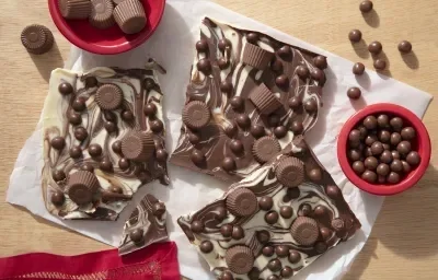 Fotografia em tons de vermelho em uma bancada de madeira com decoração de objetos vermelhos, com uma foto vista de cima. Ao centro, a barra de chocolate ao leite e branco com alguns Nescau Ball e bombons Alpino.