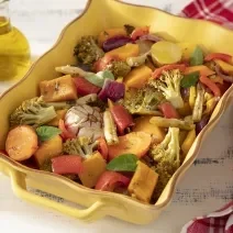 Foto da receita de Panaché de Legumes. Observa-se um refratário amarelo com os legumes cozidos. Ao lado esquerdo, um pote de vidro com azeite e, do direito, uma toalha quadriculada vermelha