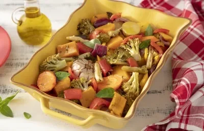 Foto da receita de Panaché de Legumes. Observa-se um refratário amarelo com os legumes cozidos. Ao lado esquerdo, um pote de vidro com azeite e, do direito, uma toalha quadriculada vermelha