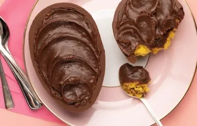 Fotografia em tons de rosa em uma bancada de madeira rosa, um pano rosa, um prato rosa redondo com o ovo de Páscoa de colher de bolo de cenoura recheado com brigadeiro.