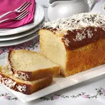 Fotografia em tons de branco de uma bancada branca com um prato retangular com m bolo. Ao fundo pratos brancos com um paninho rosa e garfos. Ao lado uma jarra e uma xícara de chá.