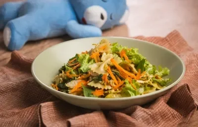 Fotografia em tons de verde e marrom em uma bancada de madeira, um prato redondo fundo verde com a salada de macarrão, frango, legumes e molho pesto dentro. Ao fundo, um ursinho azul deitado. Fotografia em tons de verde e marrom em uma bancada de madeira, um prato redondo fundo verde com a salada de macarrão, frango, legumes e molho pesto dentro. Ao fundo, um ursinho azul deitado.