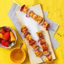Fotografia em tons de amarelo em uma bancada de madeira amarela, uma tábua de madeira, paninho azul xadrez e três espetinhos de frango. Ao lado, potinhos com pimentas e molho thailandês.