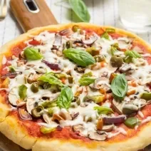 Fotografia de uma pizza com massa de pão sírio com molho de tomate, pedaços de tomate, requeijão e manjericão. A pizza está apoiada sobre uma base de madeira, que está sobre um pano de mesa branco com listras, e ao fundo, dois potinhos e um copo de vidro.