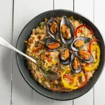 Fotografia em tons de branco e preto de uma bancada branca, sobre ele um prato preto com arroz com frutos do mar e uma colher.
