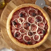 Fotografia em tons de marrom em uma bancada de madeira, uma tábua de madeira redonda com a pizza de calabresa em cima dela. Ao lado, cebola, potinho de azeite e cortador de pizza.