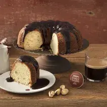 Fotografia em tons de marrom de uma bancada vista de frente, contém um pratinho redondo branco com uma fatia de bolo, um suporte para servir bolo com um bolo com cobertura e ao lado uma xicara transparente com café