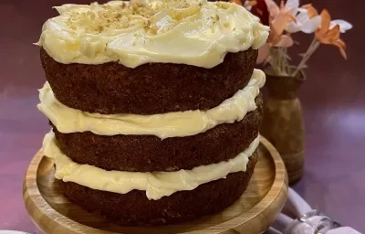 Foto em tons de marrom da receita de carrot cake servida sobre uma tábua redonda de madeira. Ao lado esquerdo há um prato de porcelana com um garfo prateado, ao lado direito há um lenço cor de rosa