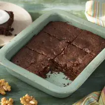 Fotografia em tons de verde em uma bancada de madeira verde, um recipiente verde quadrado com o brownie de chocolate com nozes e um pano colorido ao lado. Ao fundo, um prato redondo bege com uma fatia do brownie.