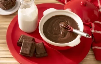 Fotografia em tons de vermelho em uma bancada de madeira de cor marrom. Ao centro, um prato redondo e vermelho com uma panela branca contendo o ganache. Ao lado, há uma garrafa de leite e ao fundo, um pano vermelho quadriculado. Fotografia em tons de vermelho em uma bancada de madeira de cor marrom. Ao centro, um prato redondo e vermelho com uma panela branca contendo o ganache. Ao lado, há uma garrafa de leite e ao fundo, um pano vermelho quadriculado.