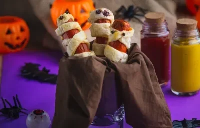 Imagem aproximada da receita de salsichas envoltas em tiras de massa folhada com pontinhos pretos, simbolizando olhos. Na cena há abóboras falsas, dois potes com ketchup e mostarda, algumas aranhas falsas, tudo numa superfície em tom roxo escuro Imagem aproximada da receita de salsichas envoltas em tiras de massa folhada com pontinhos pretos, simbolizando olhos. Na cena há abóboras falsas, dois potes com ketchup e mostarda, algumas aranhas falsas, tudo numa superfície em tom roxo escuro