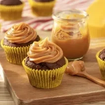 Fotografia em tons de amarelo em uma bancada de madeira de cor marrom. Ao centro, uma tábua de madeira contendo os cupcakes e um pote contendo o doce e leite com uma colher ao lado. Ao fundo, há um pano vermelho listrado.