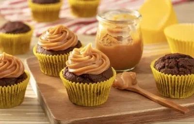 Fotografia em tons de amarelo em uma bancada de madeira de cor marrom. Ao centro, uma tábua de madeira contendo os cupcakes e um pote contendo o doce e leite com uma colher ao lado. Ao fundo, há um pano vermelho listrado.