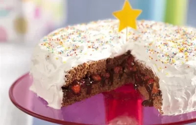 Fotografia em tons de rosa em uma bancada cinza com um suporte para bolo central na cor rosa com o bolo de aniversário confeitado em cima com cobertura branca e confeitos coloridos, cortado ao meio e recheado com chocolate e morango.