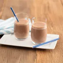 Fotografia em tons de marrom e azul de uma bancada de madeira com um prato branco, sobre ele dois copos de vidro com chocolate gelado e dois canudos azuis. Ao lado um paninho listrado branco e azul.