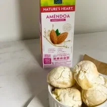 Foto da receita de pão de queijo vegano servida em diversas porções em um prato de porcelana branca sobre uma tábua de madeira com uma embalagem de nature's heart bebida de amêndoa ao fundo