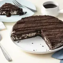 Imagem aproximada da receita de Torta de Sorvete Moça com Calda Quente, servida em um prato branco, sobre uma bancada clara decorada com um tecido azul, um pote branco com calda de chocolate e alguns utensílios