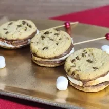 Fotografia mostra marshmallows dourados espetados entre biscoitos com cobertura de chocolate.