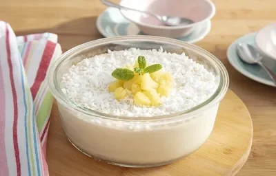 Foto em tons de branco da receita de gelado de abacaxi servida em um recipiente de vidro redondo sobre uma tábua circular de madeira com um pano colorido ao lado e xícaras brancas ao fundo