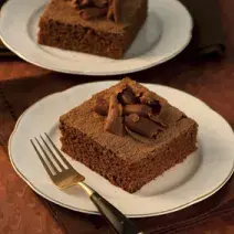 Fotografia em tons de marrom em uma mesa de madeira escura com dois pratos rasos brancos com dois pedaços do bolo de chocolate decorado com raspas de chocolate.