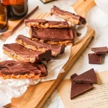 Fotografia em tons de marrom em uma bancada de madeira clara, uma tábua de madeira, barras de chocolate com pasta de amendoim ao centro e pedaços de chocolate soltos ao lado.