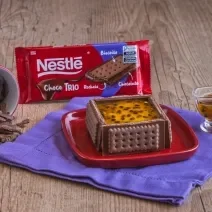 Fotografia de um bolo mousse de maracujá sobre um prato quadrado vermelho. Ao redor do bolo nota-se o chocolate choco trio, além de uma calda de maracujá por cima do bolo.