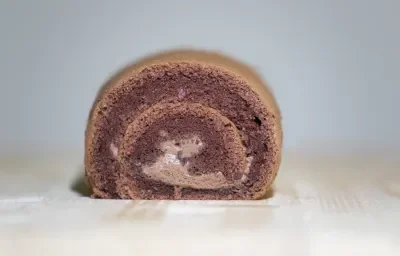 Fotografia de um bolo de rolo de chocolate, em tons de marrom, ao centro de uma mesa de madeira clara.