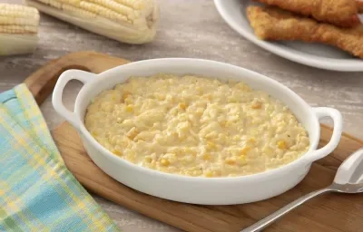 Foto da receita de Creme de Milho Cremoso. Observa-se um refratário branco com o creme de milho, do lado esquerdo superior espigas e do lado direito superior filés de frango empanados. O refratário do creme está sobre uma tábua de madeira. Foto da receita de Creme de Milho Cremoso. Observa-se um refratário branco com o creme de milho, do lado esquerdo superior espigas e do lado direito superior filés de frango empanados. O refratário do creme está sobre uma tábua de madeira.
