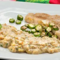 Fotografia em tons de verde em uma mesa com uma toalha verde florida, um prato branco grande com o peixe servido com quiabo e com purê de banana da terra.