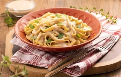 Fotos da receita de macarrão ao molho branco servido em um prato adornado laranja sobre um pano quadriculado vermelho e branco com talheres com cabo de madeira ao lado direito. Ao fundo uma taça de vidro com o molho branco. Fotos da receita de macarrão ao molho branco servido em um prato adornado laranja sobre um pano quadriculado vermelho e branco com talheres com cabo de madeira ao lado direito. Ao fundo uma taça de vidro com o molho branco.