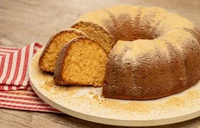 Foto de uma bancada de madeira. Sobre ela há um tabuleiro branco com um bolo polvilhado de Farina Láctea Nestlé e com duas fatias cortadas. Ao lado do tabuleiro há um tecido listrado vermelho e branco