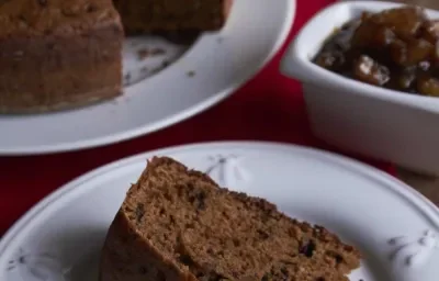 Fotografia em tons de marrom em uma mesa de madeira com um guardanapo vermelho, um prato branco redondo raso com uma fatia de bolo de chocolate com geleia de banana. Ao fundo, um prato maior com o bolo inteiro.