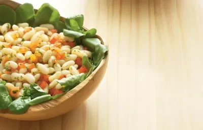 Fotografia de uma mesa de madeira com um bowl de madeira em cima com a salada dentro com folhas, feijão e cenoura.