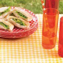 Fotografia em tons de vermelho e amarelo em uma mesa com uma toalha amarela xadrez, pratos vermelhos com bolinhas brancas e os sanduíches de presunto em cima deles. Mais à frente, copos compridos laranjas com suco e canudos de papel.